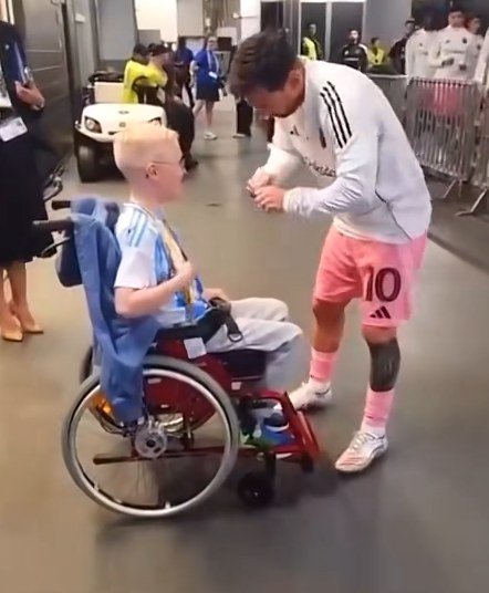 Lionel Messi se agacha para saludar a un niño en silla de ruedas que lleva la camiseta de la Selección Argentina, en el pasillo de un estadio.